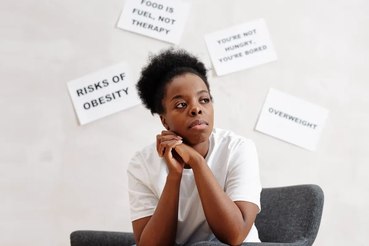 Woman in white t-shirt sits thoughtfully with posters on health and obesity around her.
