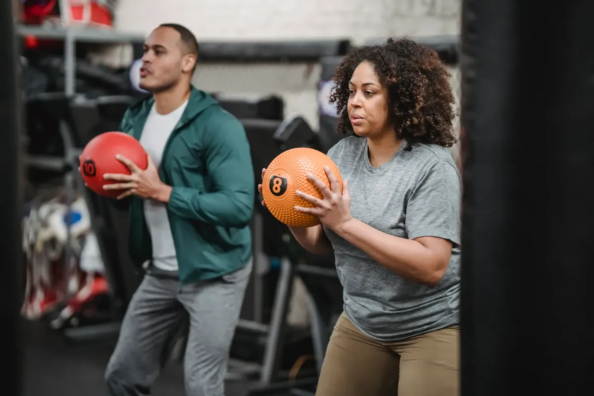 Determined young ethnic male trainer and African American female client in activewear doing exercise with medicine balls during workout in modern gym